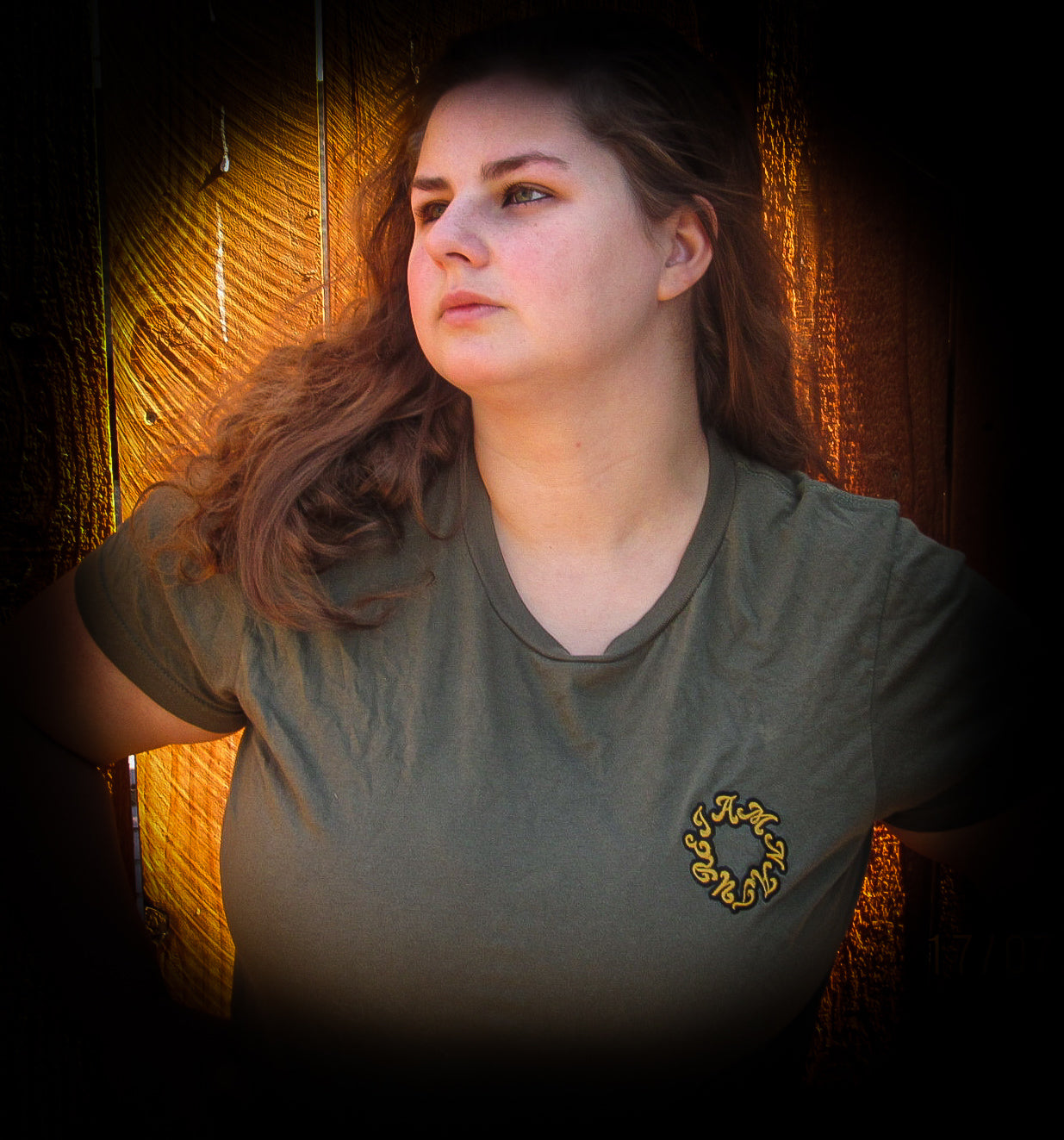 Woman wearing a military green t-shirt with a sunflower embroidered logo against a fence background